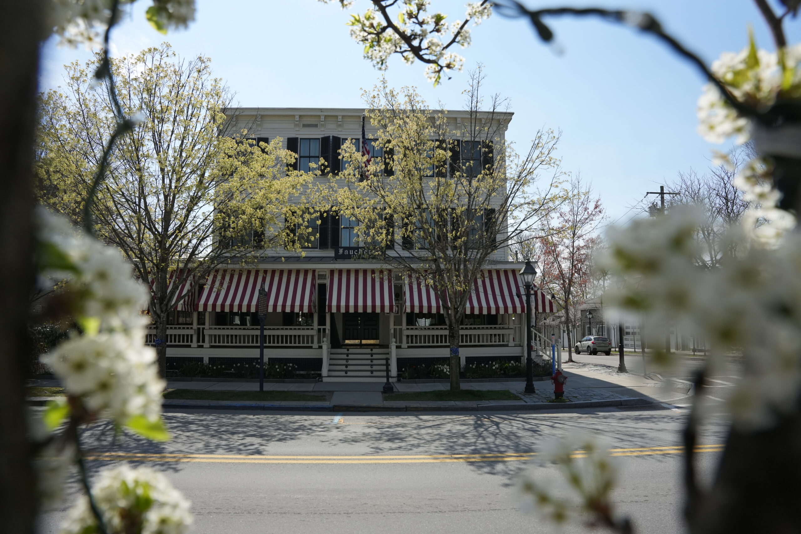 Two-story white storefront with red-and-white striped awning, framed by blooming trees and a clear blue sky, an American flag visible