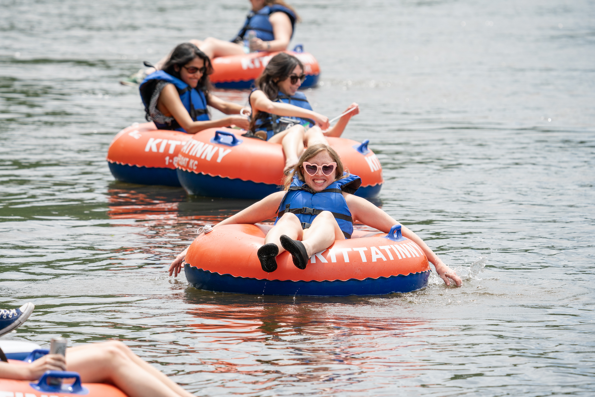 Smiling woman in a blue life jacket rides an orange-and-blue inflatable tube on a lake, with others behind her in life jackets.