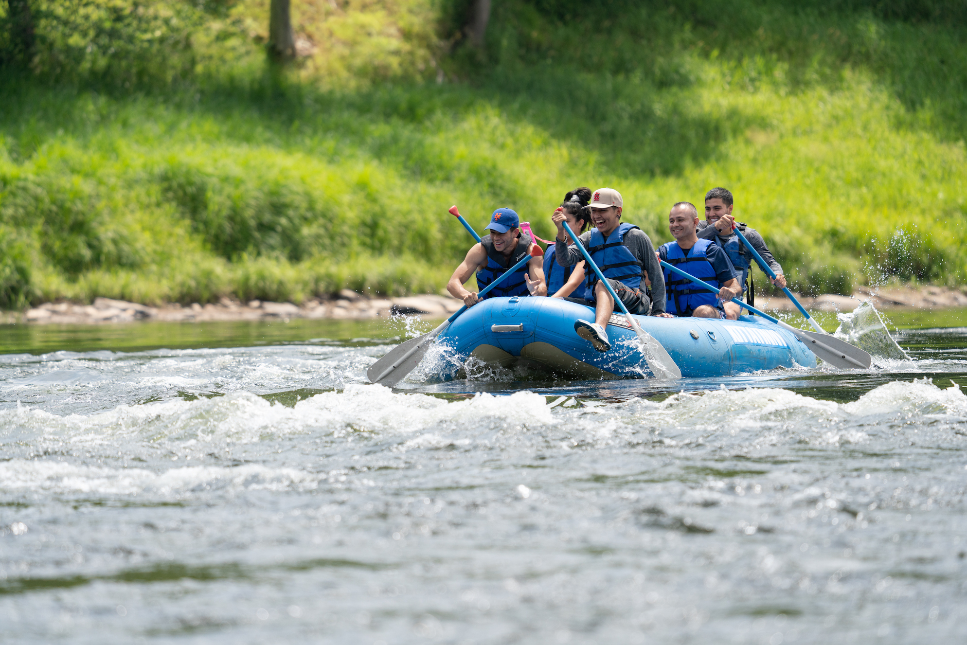 Group of friends wearing blue life jackets rafting on a river with green banks ahead of them.