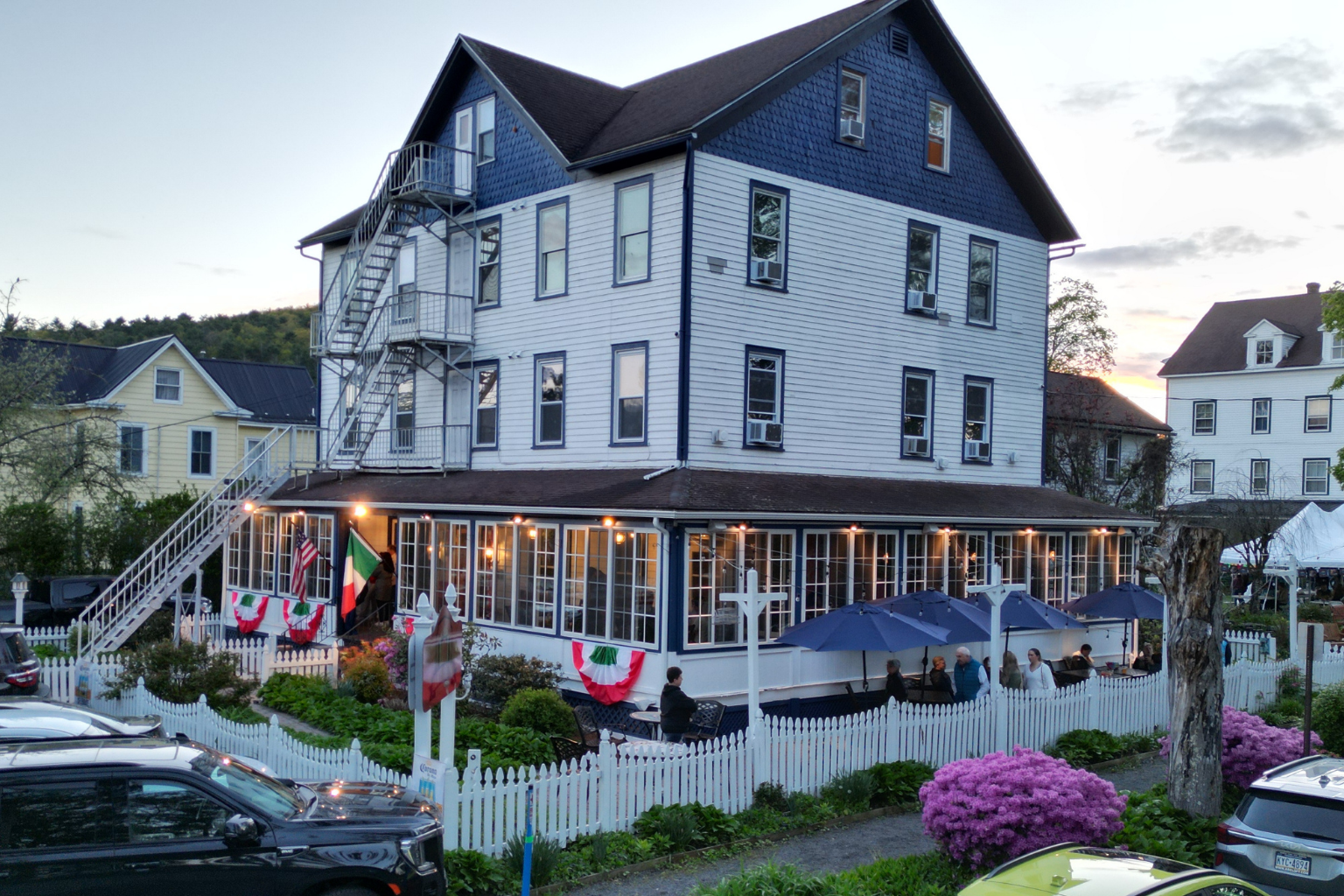Three-story white building with blue trim housing a restaurant, outdoor seating under blue umbrellas, and a white picket fence with flags at dusk.